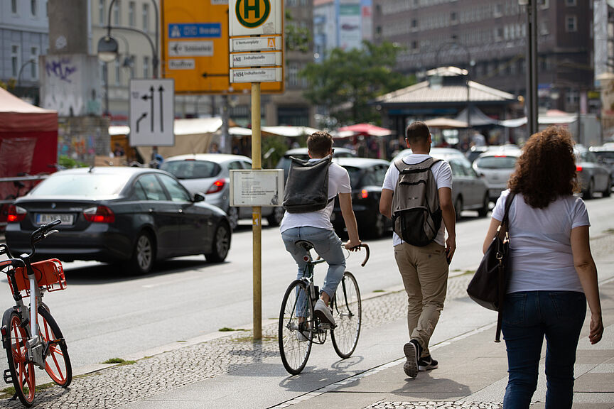 Geteilter Verkehrsraum in der Stadt Eine Straßenszene: Ein Fahrradfahrer und zwei Fußgänger teilen sich einen gepflasterten Gehweg. Im Hintergrund parken Autos am Straßenrand.
