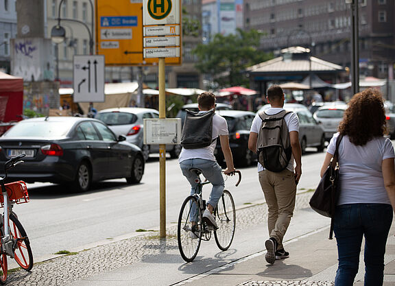 Eine Straßenszene: Ein Fahrradfahrer und zwei Fußgänger teilen sich einen gepflasterten Gehweg. Im Hintergrund parken Autos am Straßenrand.
