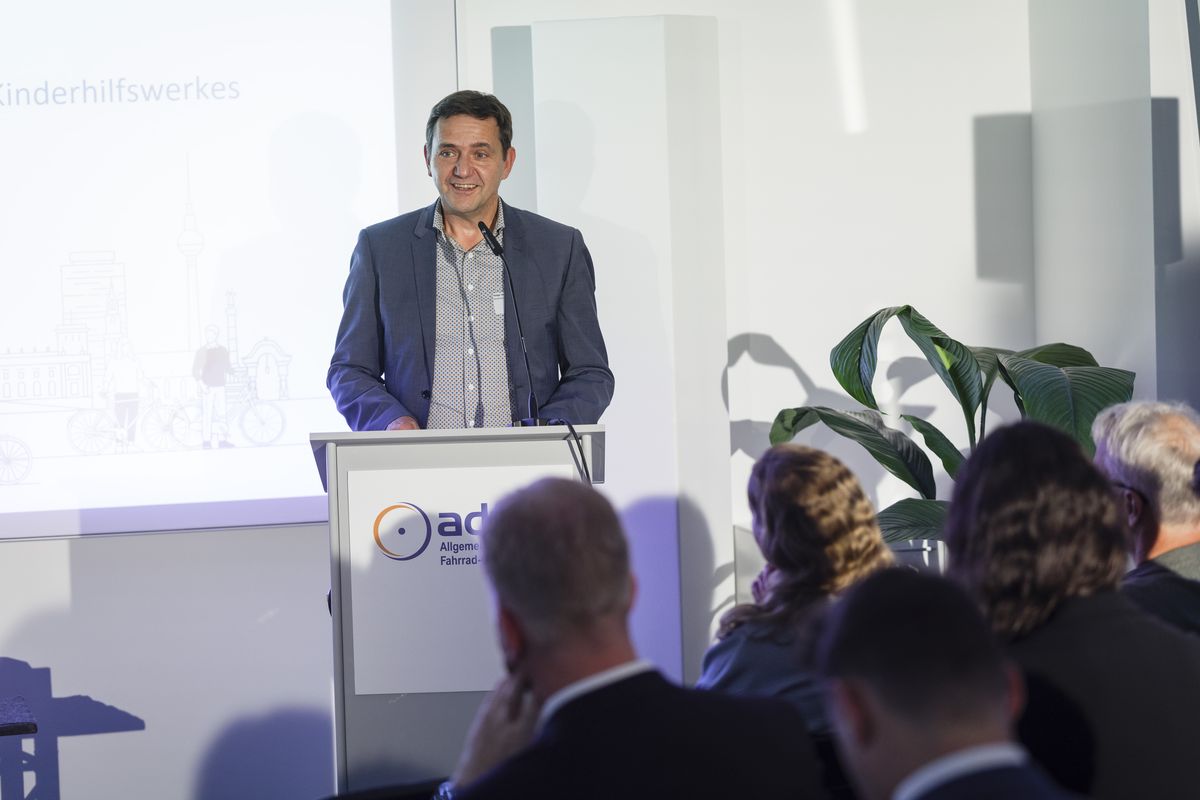 Holger Hofmann, Bundesgeschäftsführer des Deutschen Kinderhilfswerkes, am Podium. Im Hintergrund eine Leinwand, auf die Licht projiziert wird. 